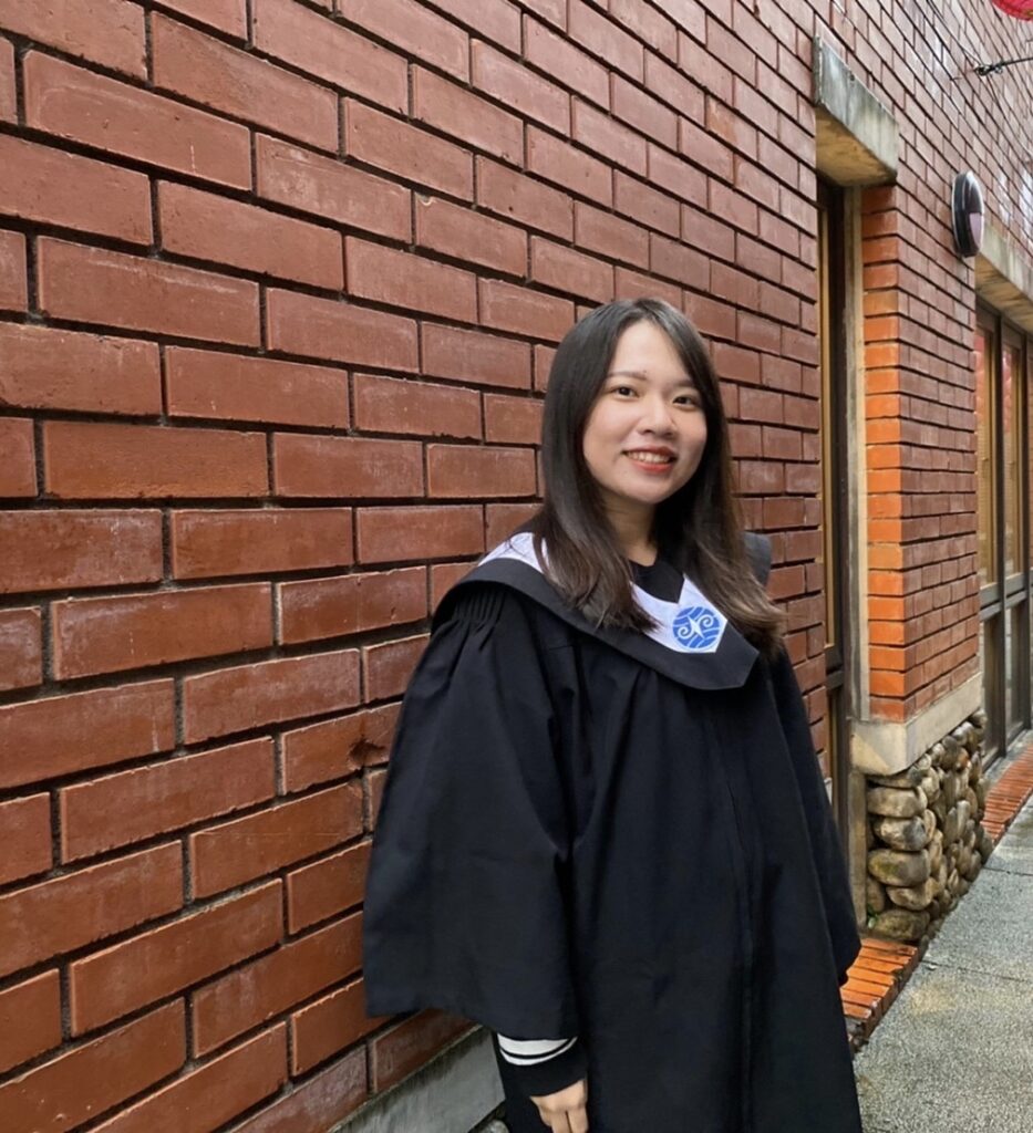 Woman in black graduation gown standing against a brick wall