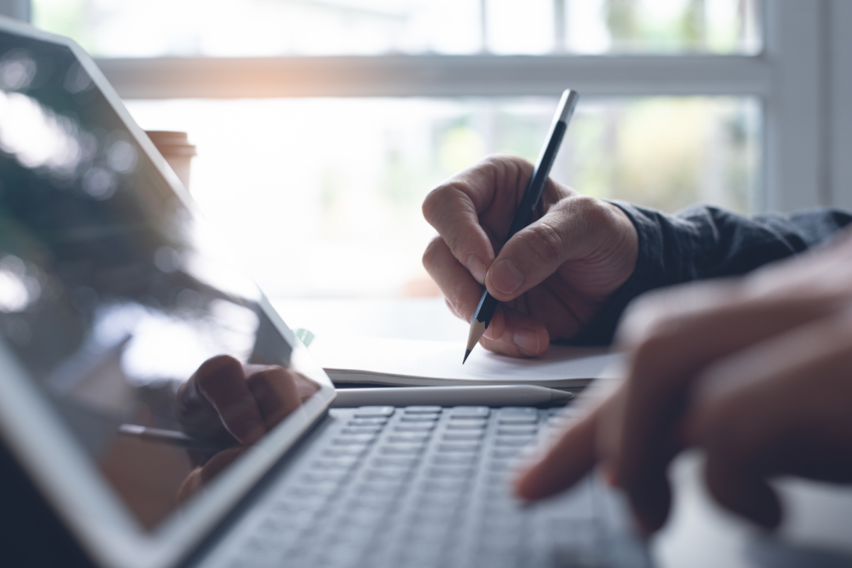 Person's hand on the laptop keyboard and their other hand writing on a paper to the side of the laptop