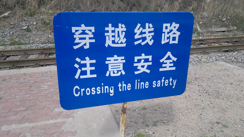 A blue sign in front of train-tracks. The sign includes Chinese and English text warning about crossing the tracks safely.
